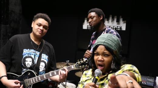 Guitarist, drummer, and singer pointing at the camera in a black-walled rehearsal room
